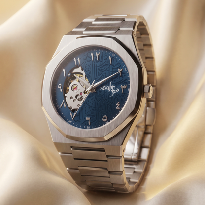 A luxury men's watch with a brushed stainless steel octagonal case and link bracelet, featuring a deep blue dial with Arabic numerals and a skeletonized side window showing the inner movement, set against draped champagne-colored fabric.